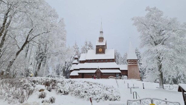 En snötäckt byggnad med ett klocktorn, omgiven av träd och buskar.