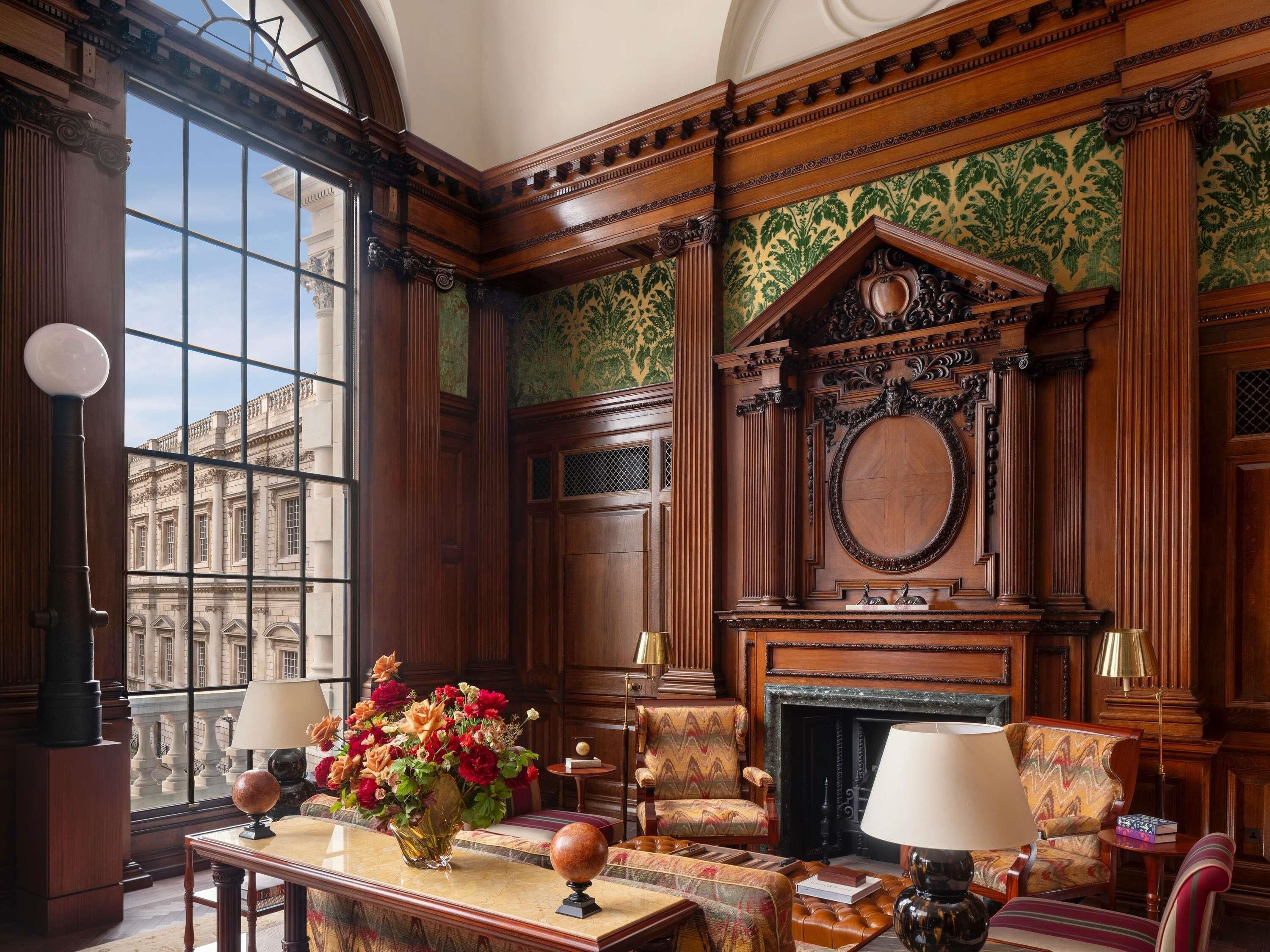 A room with a fireplace, ornate wood paneling, a large window with a view of a building, and a table with a vase of flowers.