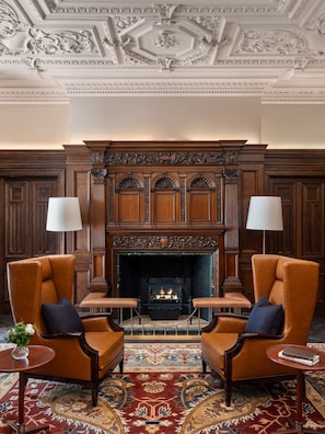 A room with a fireplace, two armchairs, a rug, and a wooden mantelpiece. - Raffles London at The OWO (London)