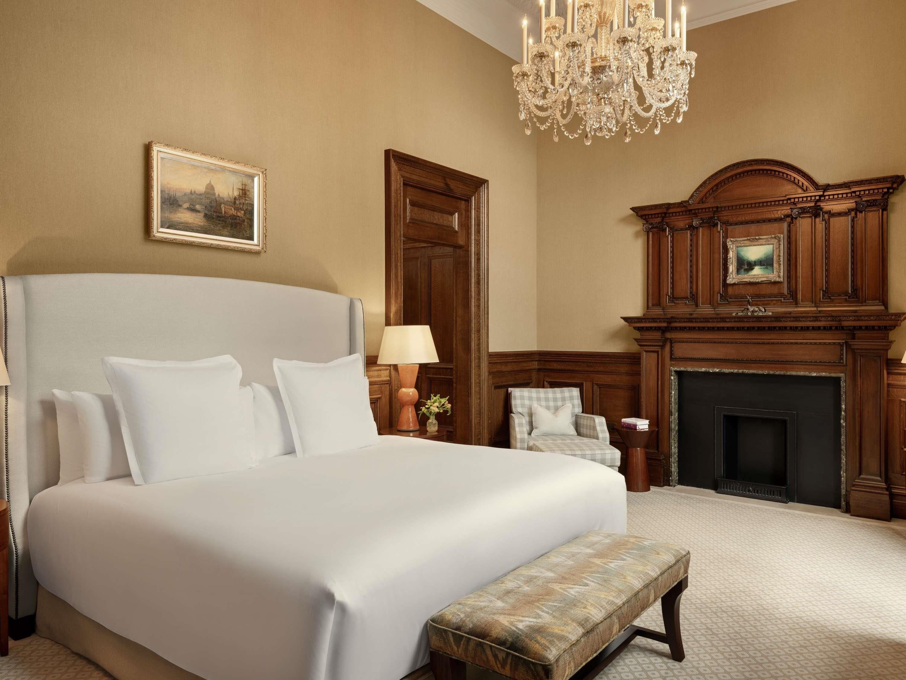 A large bed with white linens, a chandelier, a fireplace, and a painting on the wall.