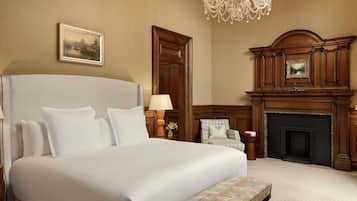 A large bed with white linens, a chandelier, a fireplace, and a painting on the wall.