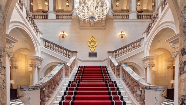 A grand staircase with red carpet, marble balustrades, and a large chandelier. - Raffles London at The OWO (London)