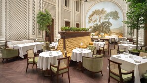 A restaurant with a high ceiling and glass panels, featuring tables and chairs arranged for dining. - Raffles London at The OWO (London)