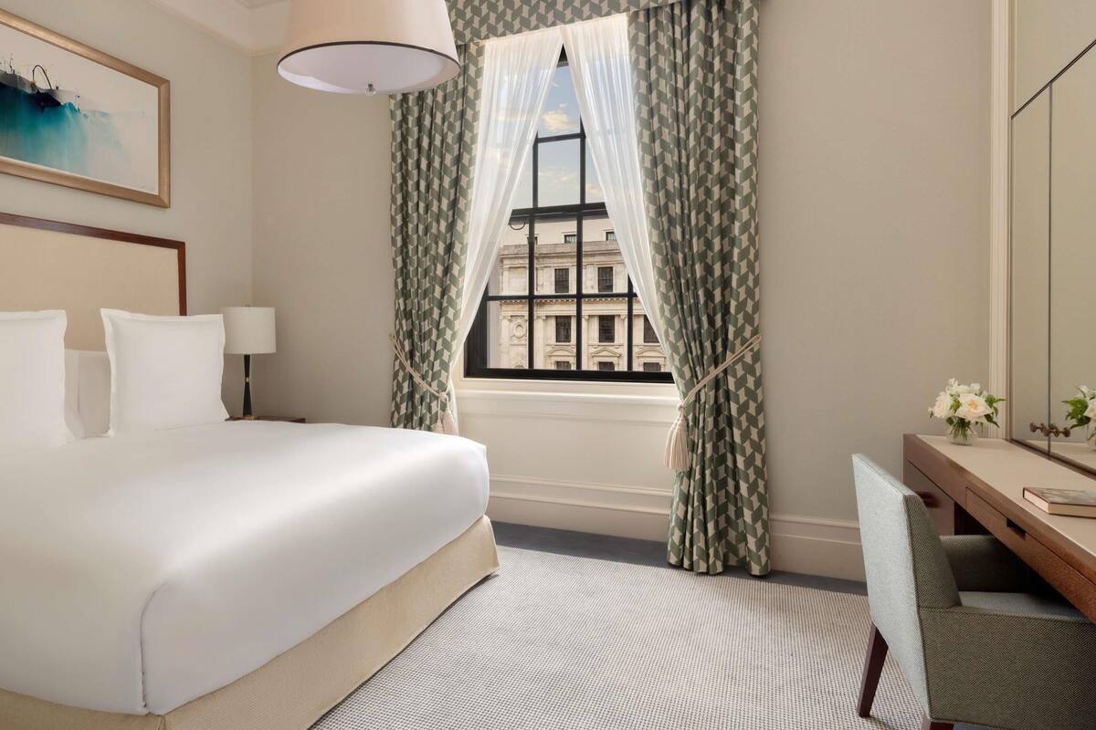 A hotel room with a bed, a desk, a chair, a window with patterned curtains, and a painting on the wall.