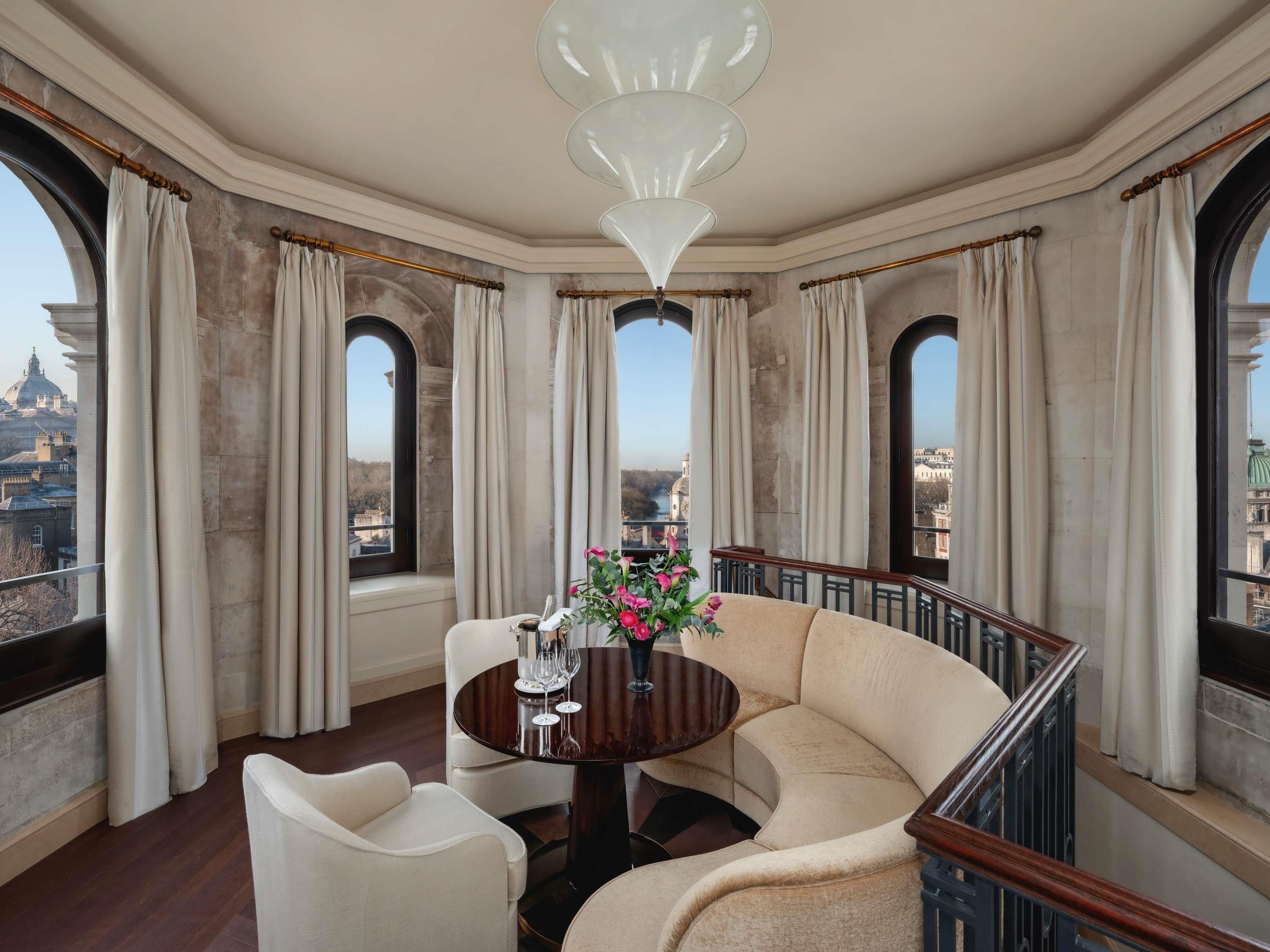 A room with a round table, two chairs, a chandelier, and large windows with curtains.
