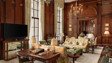 A room with a wooden paneled wall, a large window, a chandelier, a sofa set, a desk with a lamp, and a television.