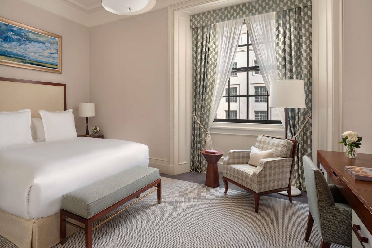 A hotel room with a large bed, a desk with a chair, a small side table, and a window with patterned curtains.