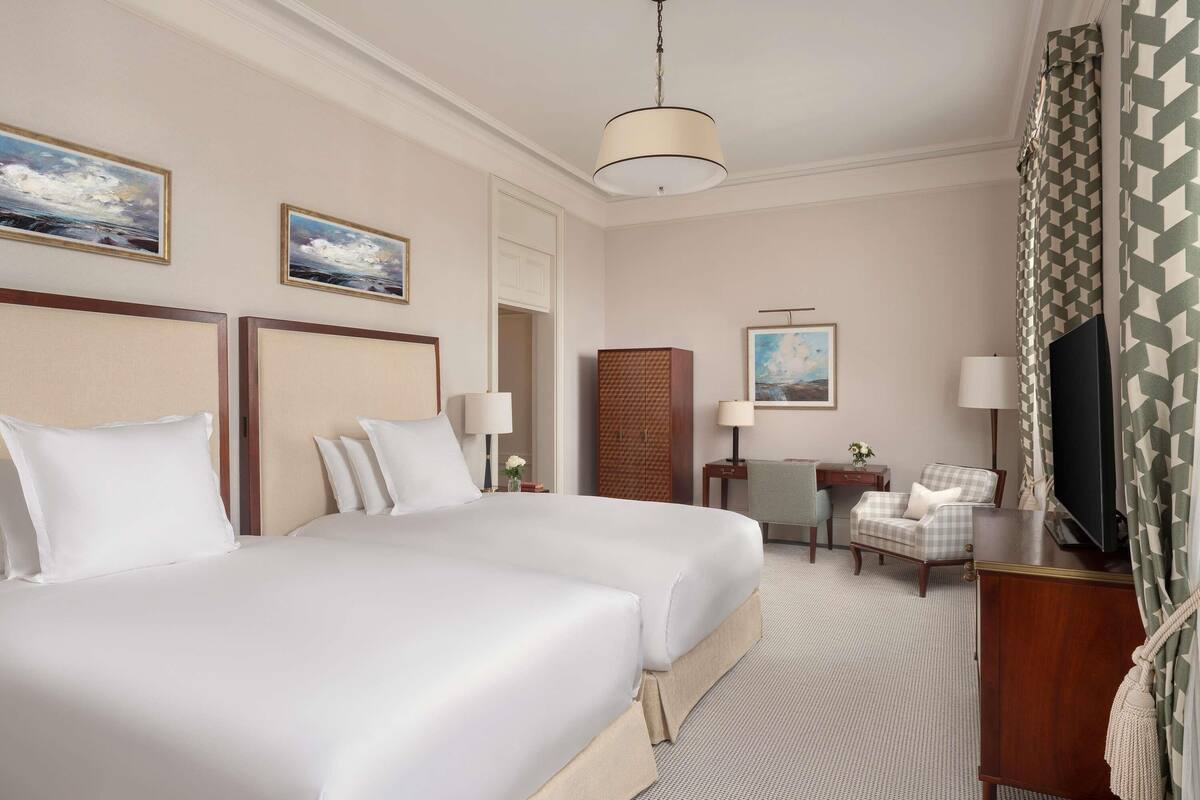 A hotel room with two beds, a desk, a chair, a TV, and artwork on the walls.