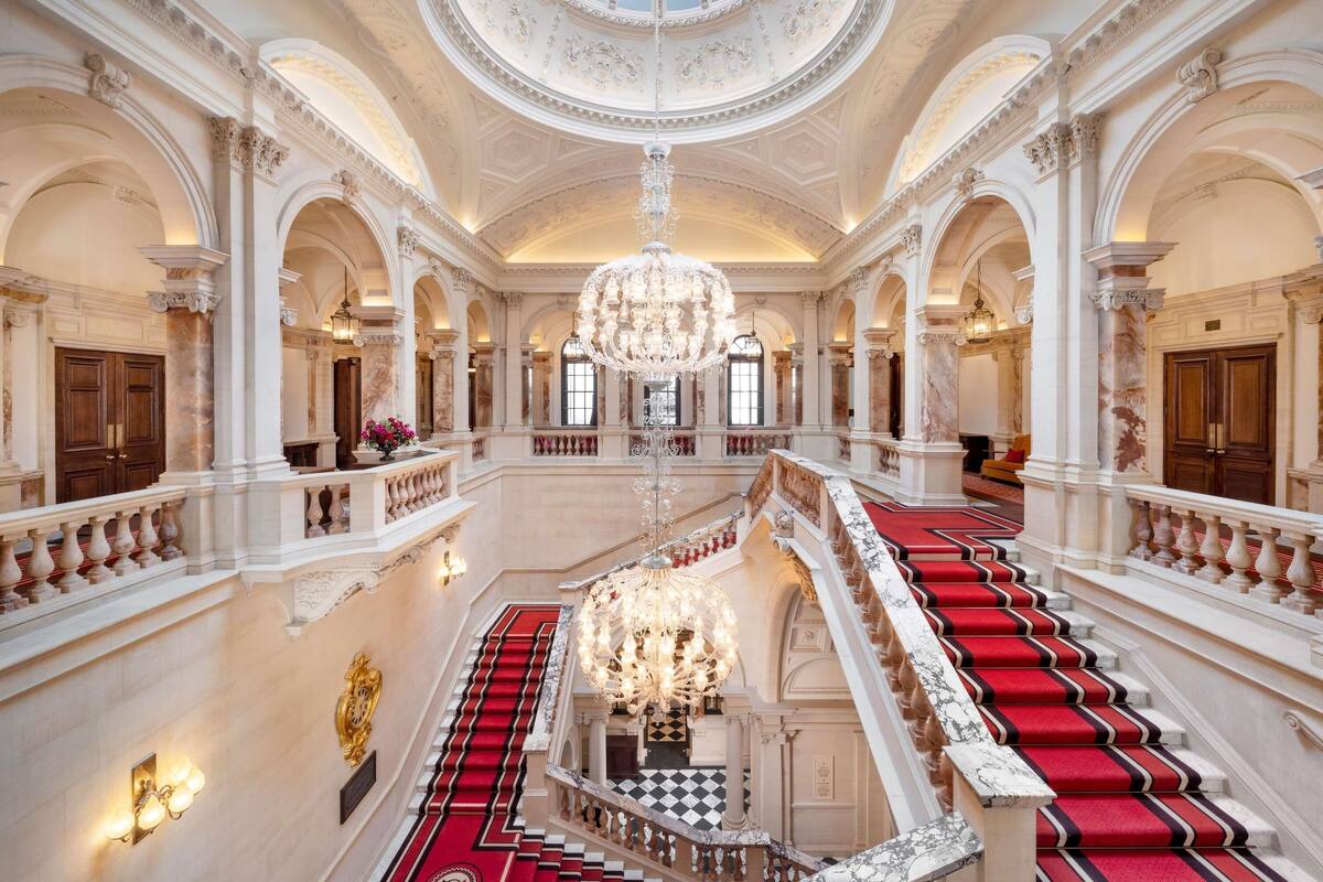 A grand interior with a central staircase, ornate columns, and a large chandelier.