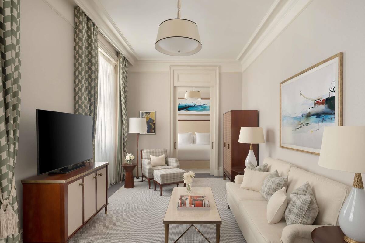 A hotel room with a flat-screen TV, a sofa, a coffee table, a cabinet, a lamp, and a painting on the wall.