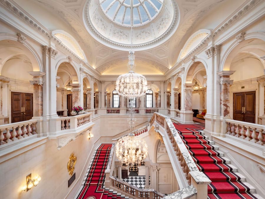 A grand interior with a central staircase, ornate columns, and a large chandelier.