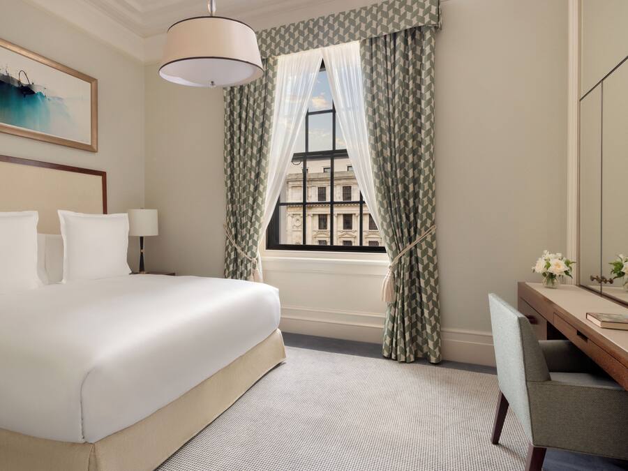 Quarto de hotel com uma cama grande, uma escrivaninha com cadeira, janela com cortinas e um quadro na parede.