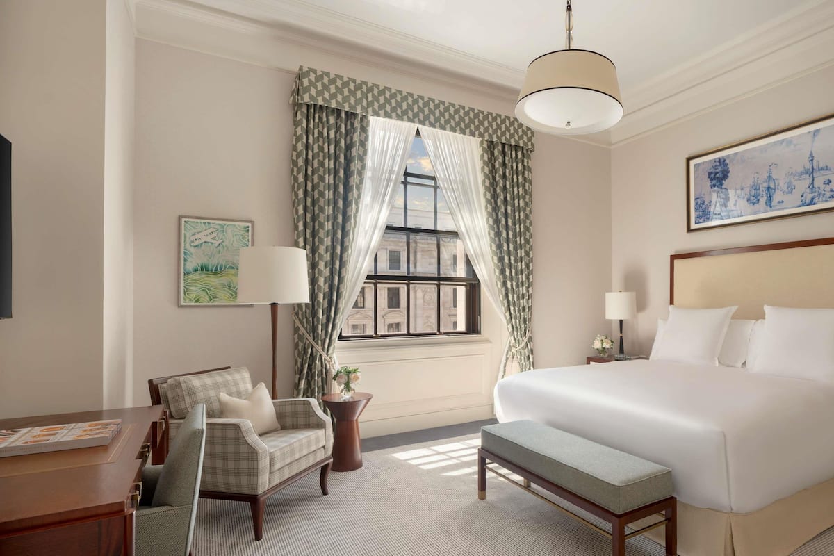 A hotel room with a bed, a desk, a chair, a lamp, a window with curtains, and a painting on the wall.
