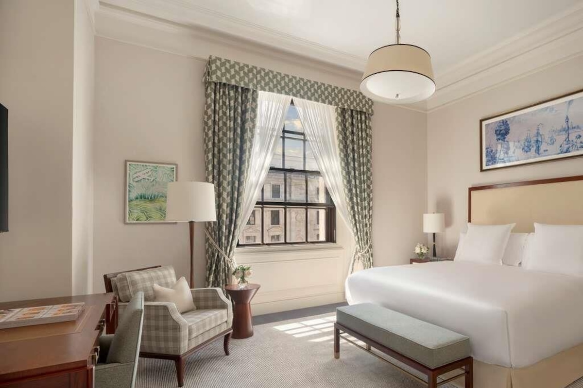 A hotel room with a large bed, a desk with a chair, a small table with a lamp, a window with curtains, and a painting on the wall.