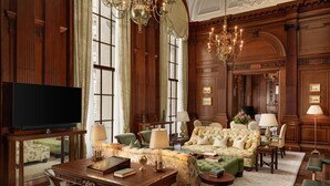 A room with a wooden paneled wall, a large window, a chandelier, a sofa set, a desk with a lamp, and a television. - Raffles London at The OWO (London)