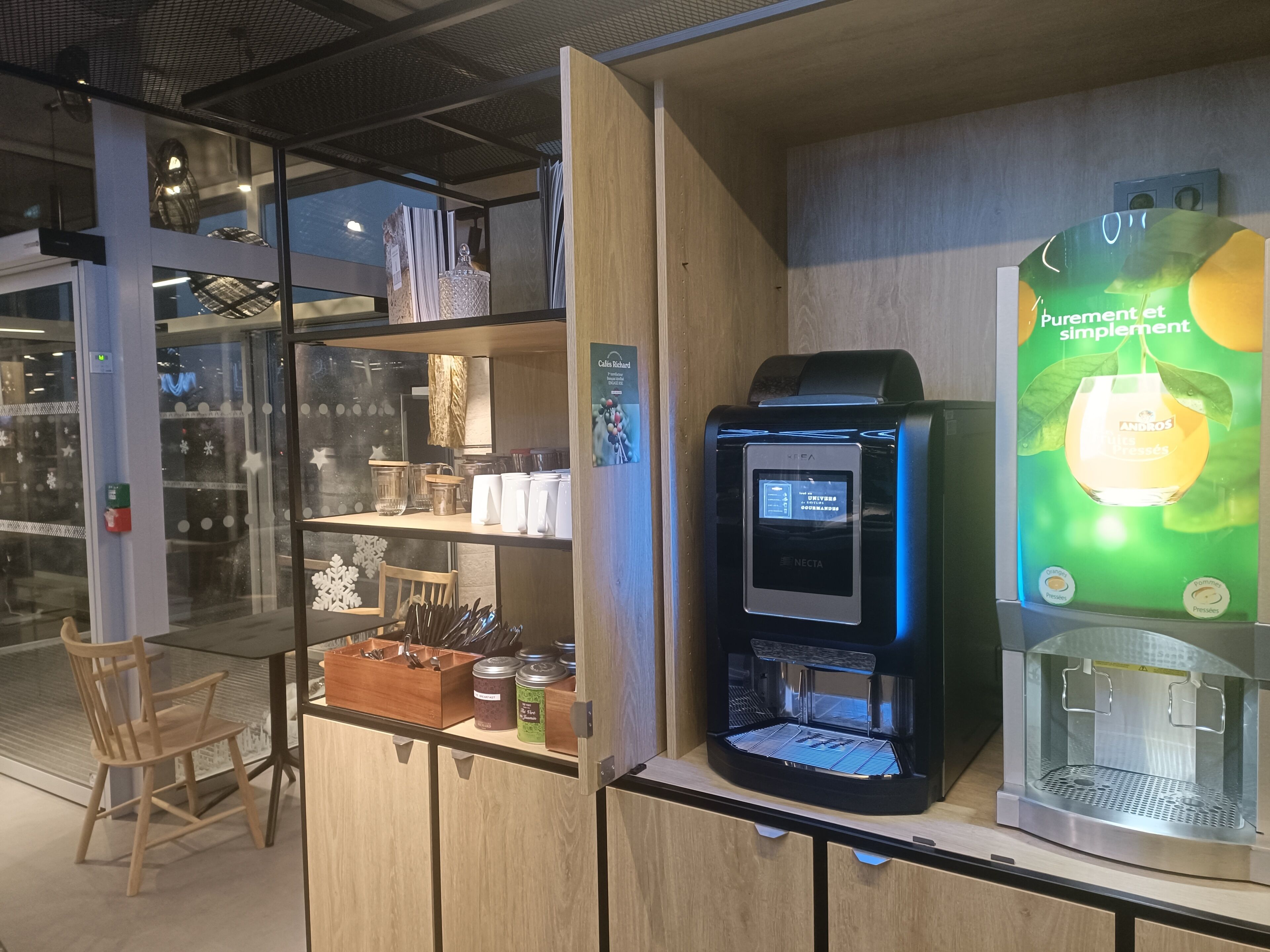 Une machine à café dotée d’un écran numérique, un présentoir en verre contenant des bocaux, et une chaise en bois.
