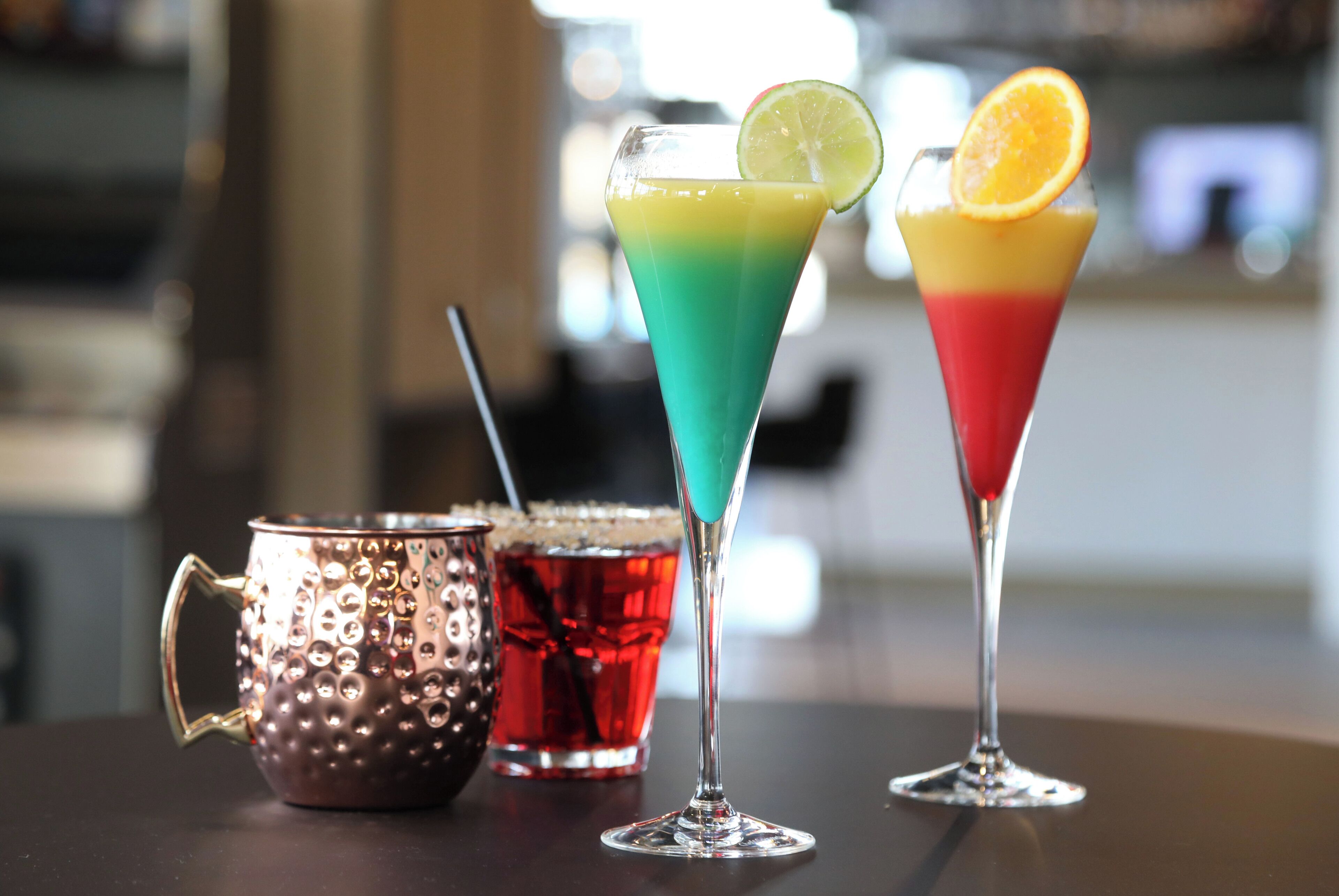 Two colorful cocktails in martini glasses with garnishes, a copper mug, and a glass of red beverage on a table.