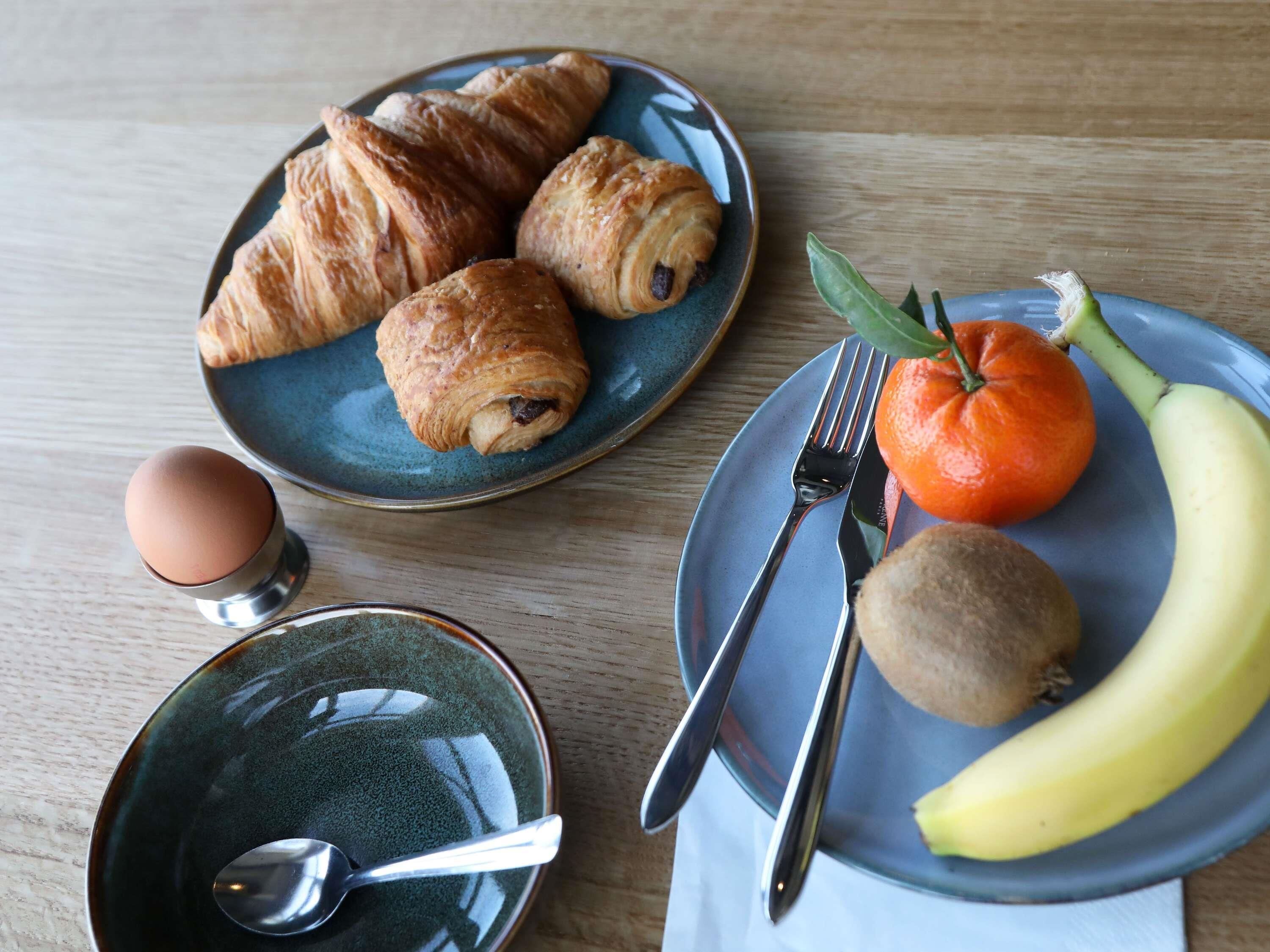 A breakfast setting with croissants, an orange, a banana, a kiwi, and a bowl of eggs on a wooden table.