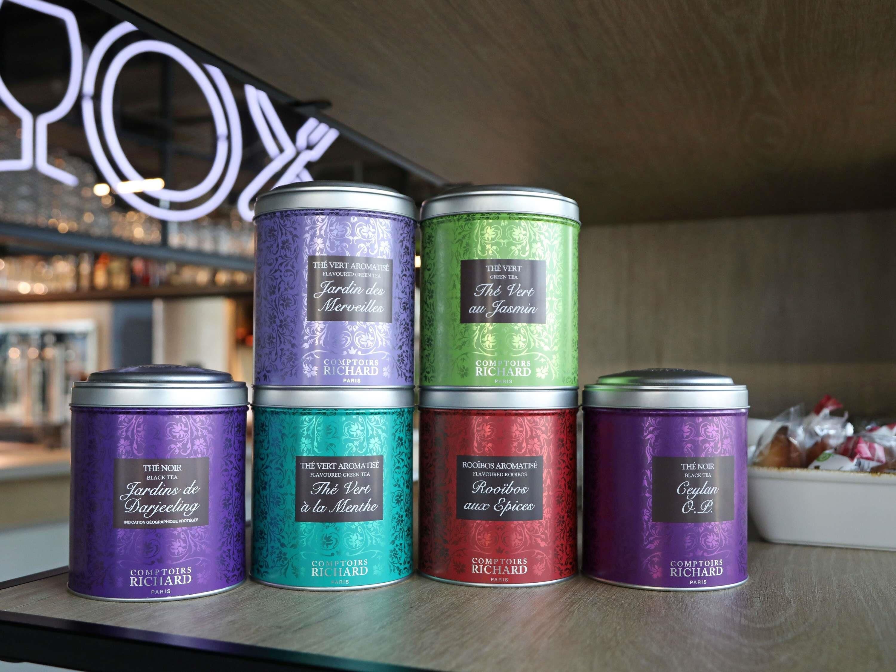 Quatre boîtes à thé de différentes saveurs, portant des étiquettes différentes, disposées sur une étagère.