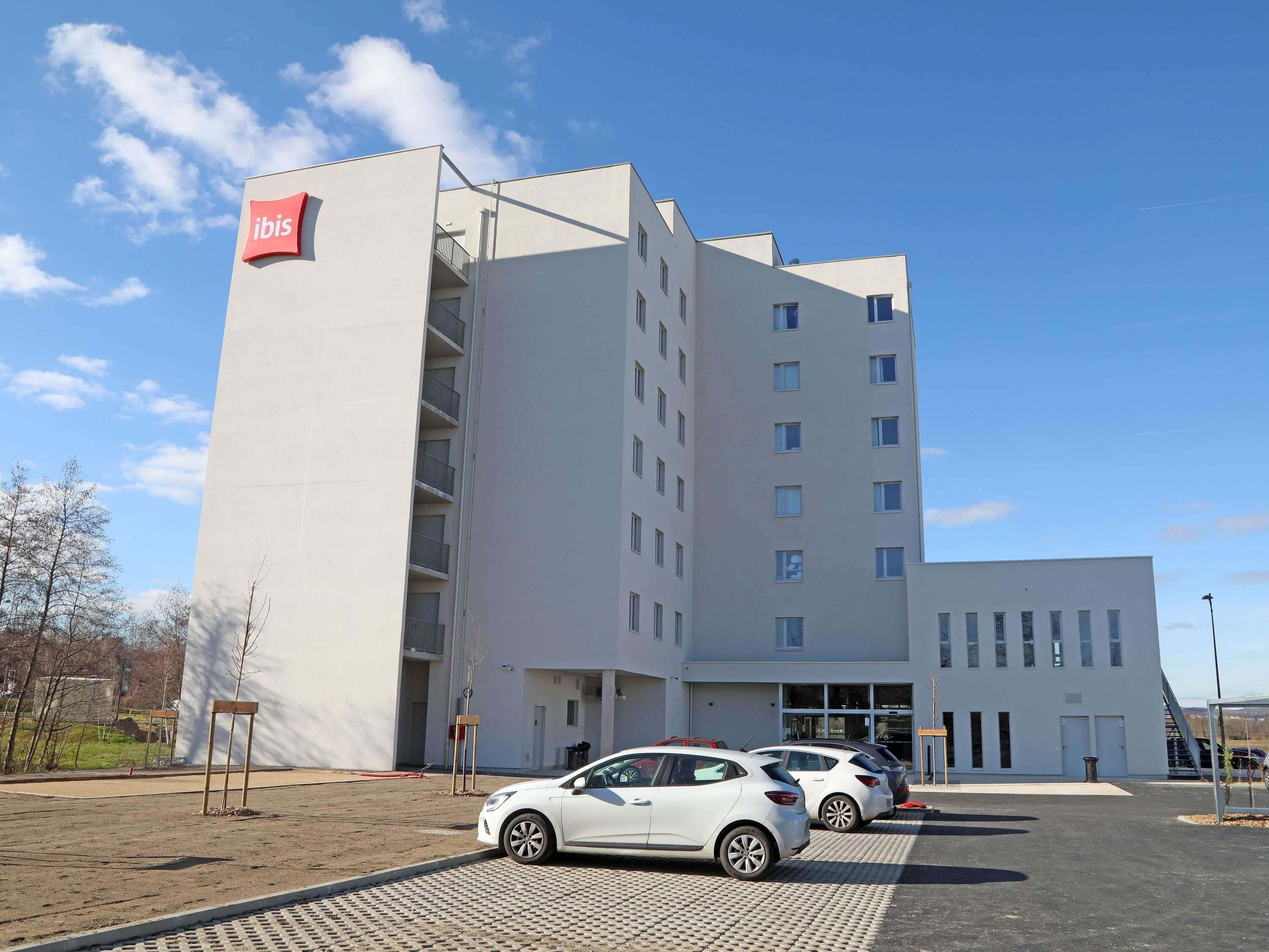 Un edificio moderno con un llamativo letrero de 'ibis', un estacionamiento con varios coches y un cielo despejado.