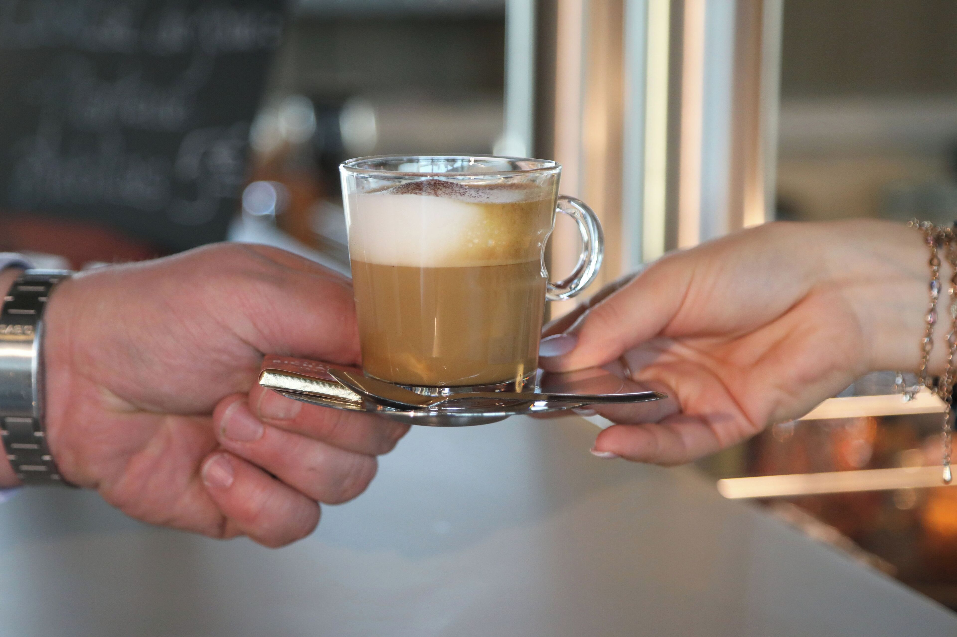 Una persona sosteniendo una taza de vidrio con una bebida, probablemente café, y una cuchara en su interior.