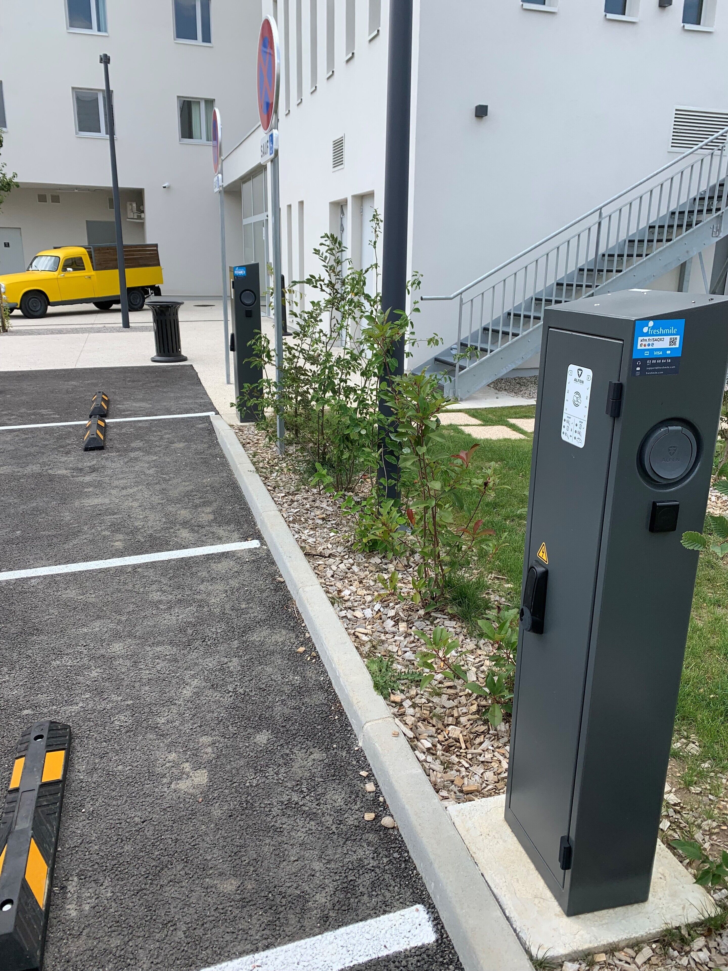Pysäköintialue, jossa on keltainen kuorma-auto, huoltorasia ja portaikko, joka johtaa rakennukseen.