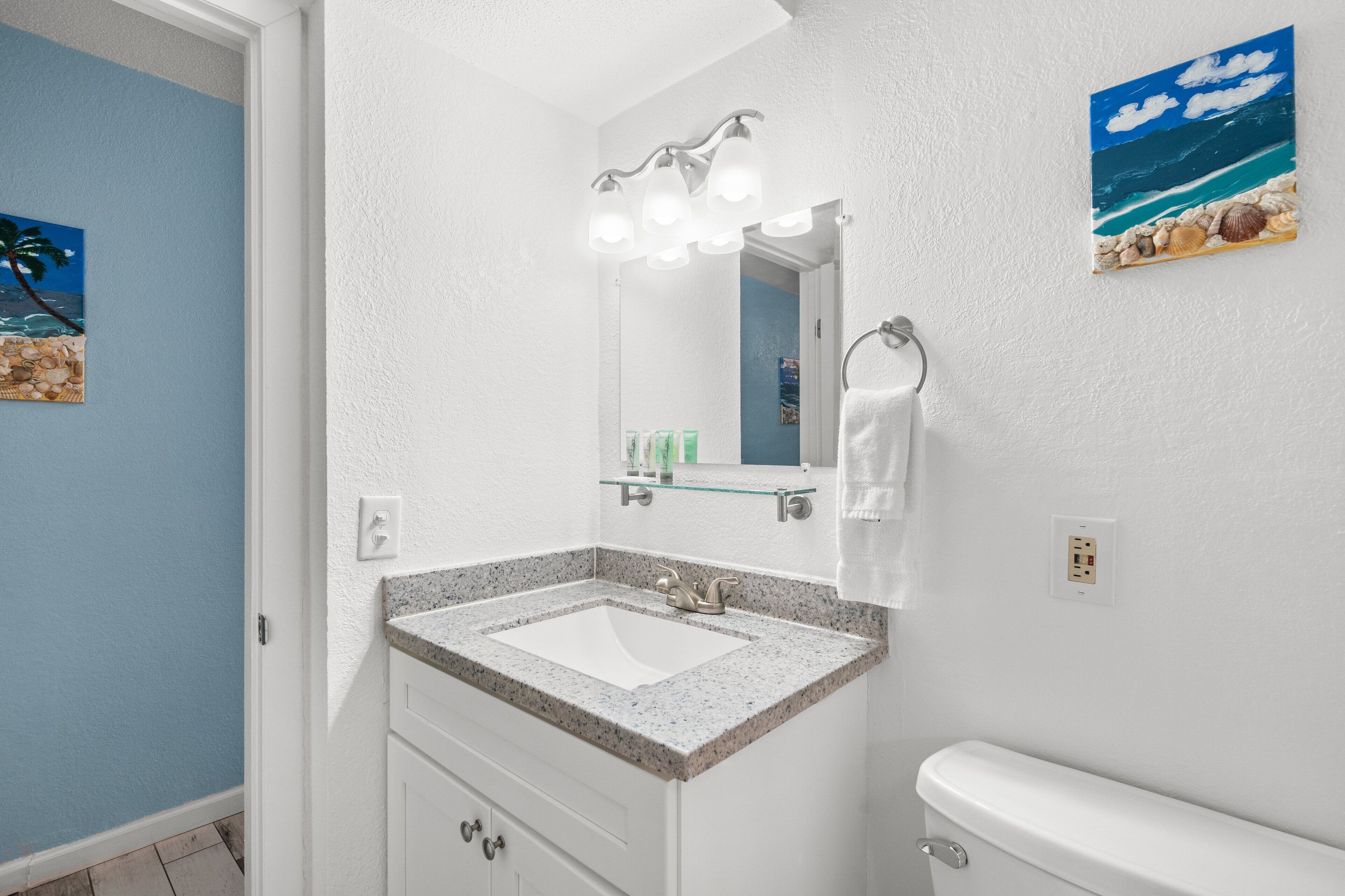 Un baño con un lavamanos blanco, un estante de vidrio arriba, un espejo con luces y una pintura de una playa en la pared.