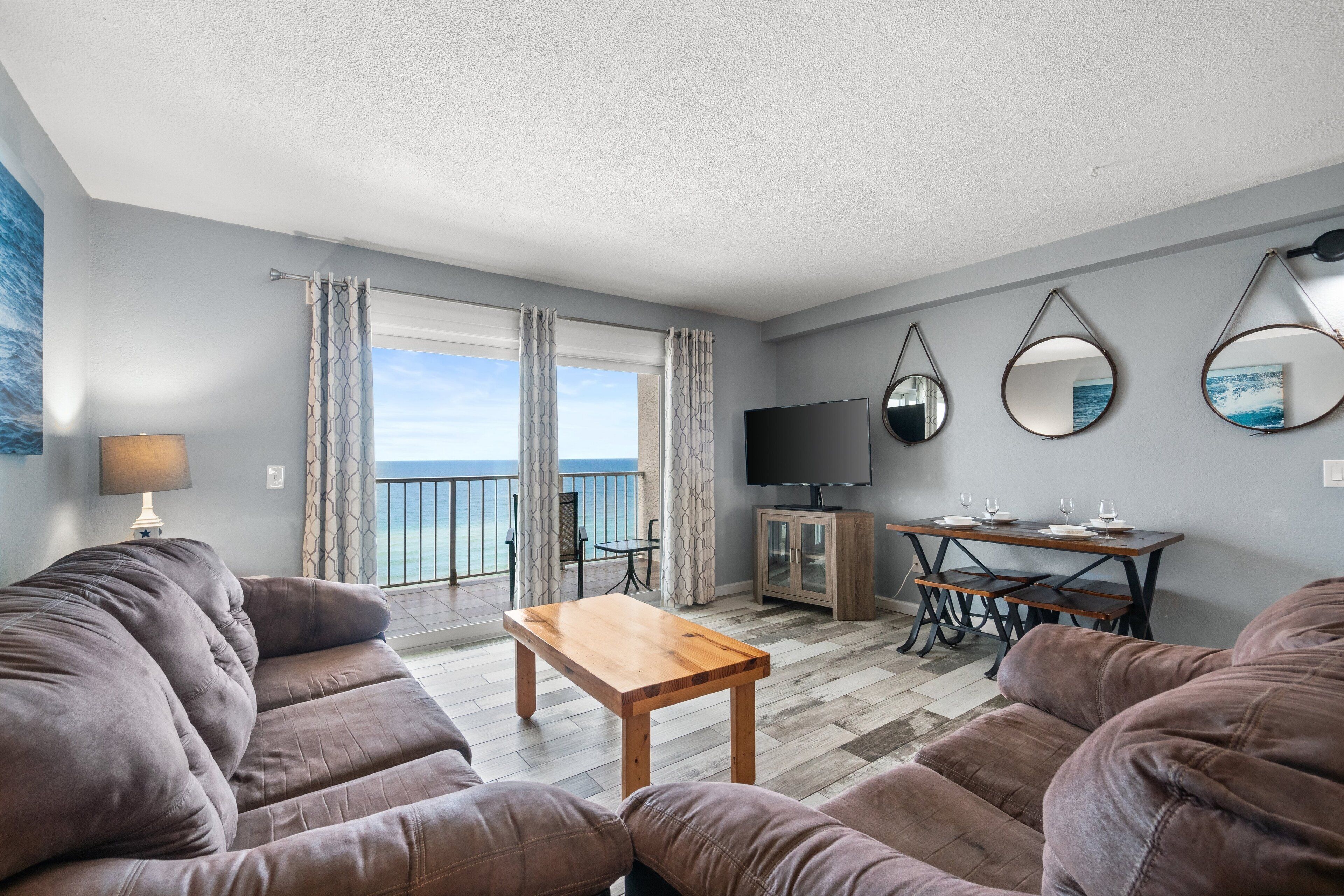 Una sala de estar con un sofá, una mesa de centro de madera, un televisor y una mesa de comedor con sillas. Hay un balcón con cortinas y vista al mar.