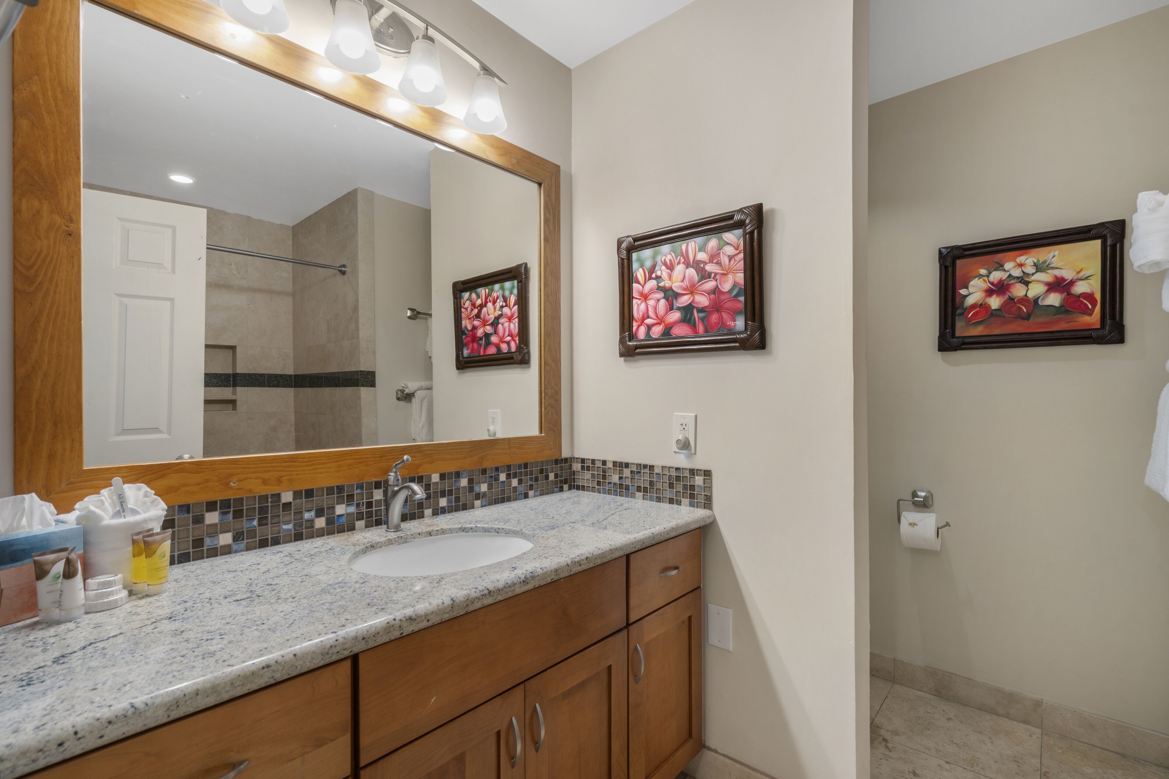 Une salle de bain avec un grand miroir, un lavabo, des meubles en bois et deux tableaux représentant des motifs floraux, encadrés et accrochés au mur.