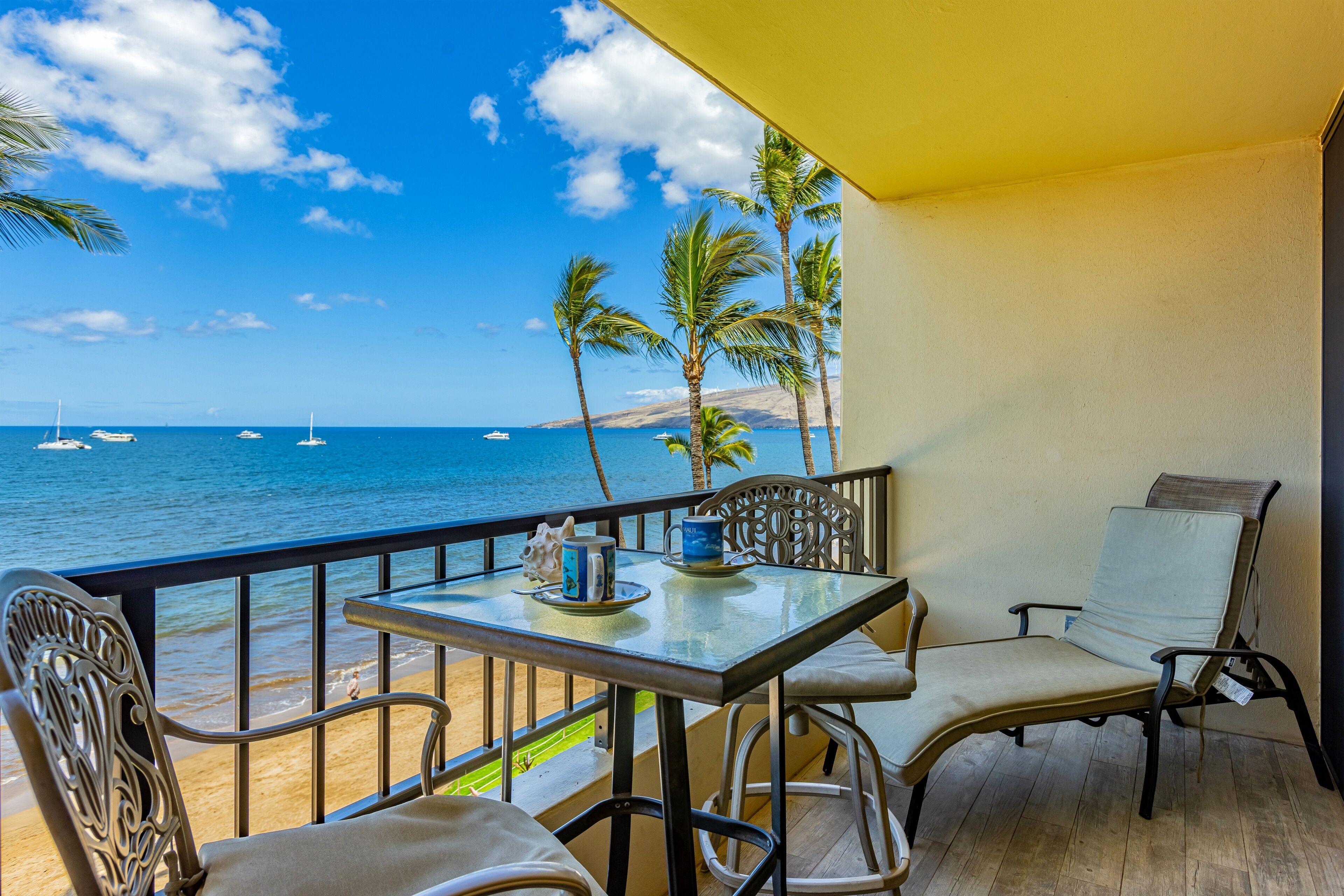 A balcony with a table and chairs overlooking a beach with boats and palm trees.