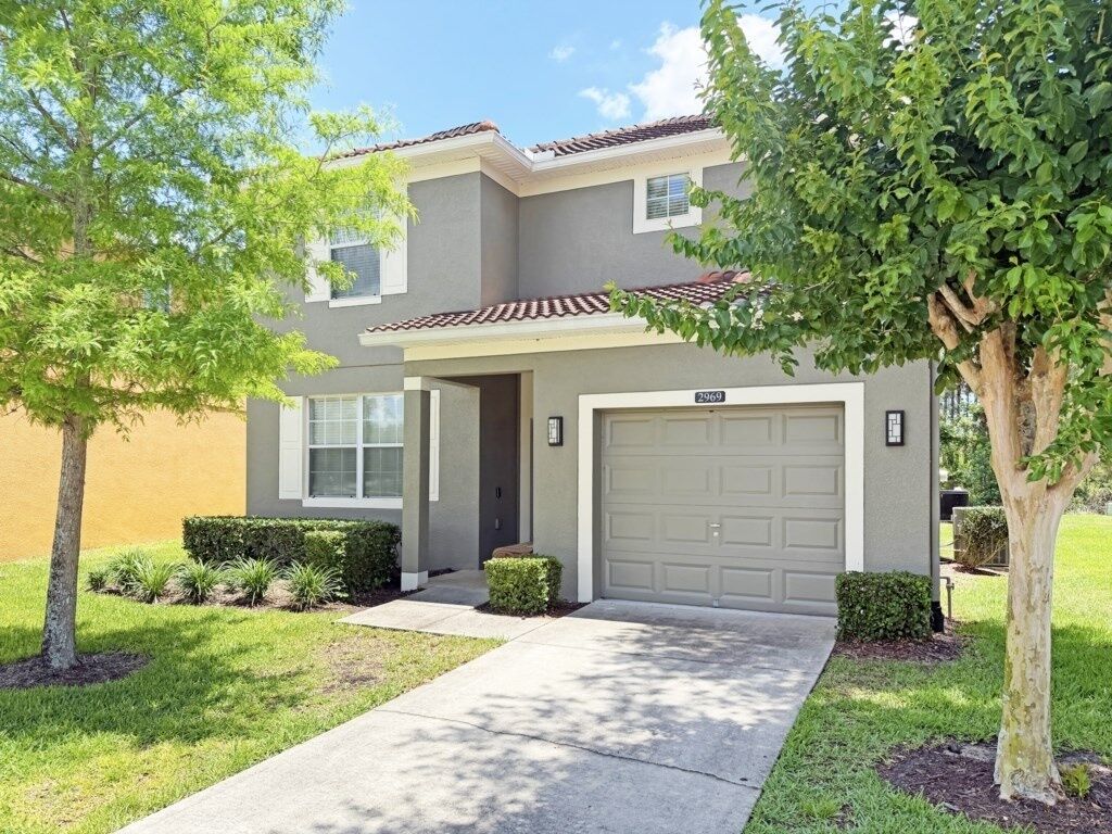 A two-story house with a garage, a front door, and a small front yard with a tree and shrubs.