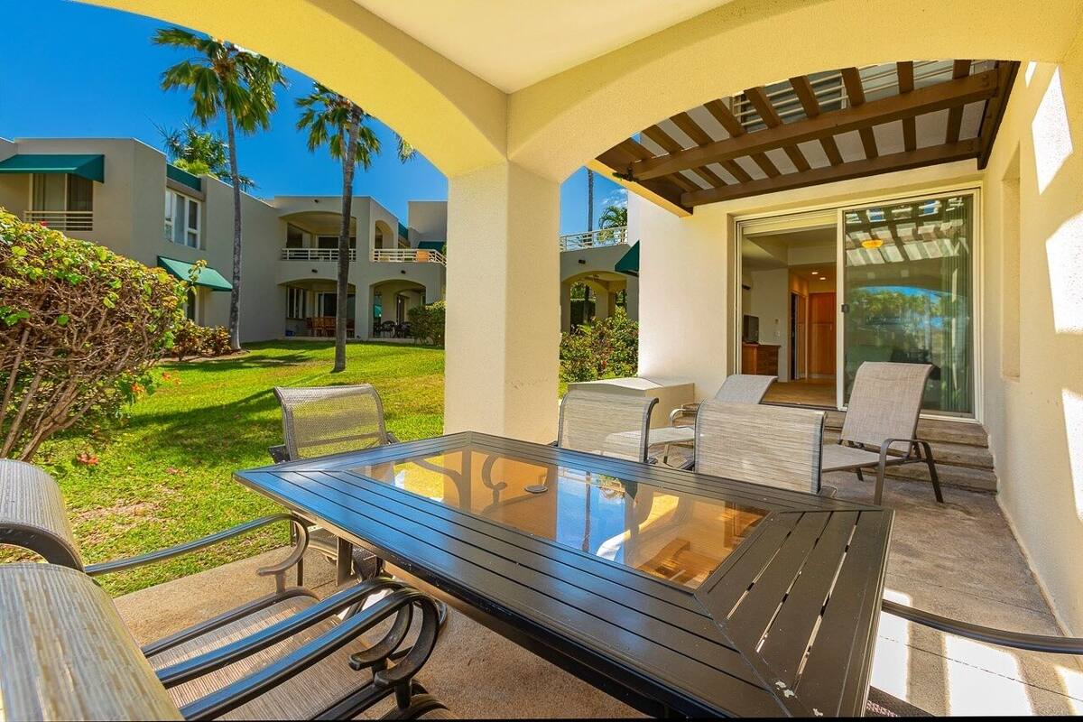 A covered outdoor seating area with a glass table and chairs, surrounded by greenery and a view of hotel buildings.