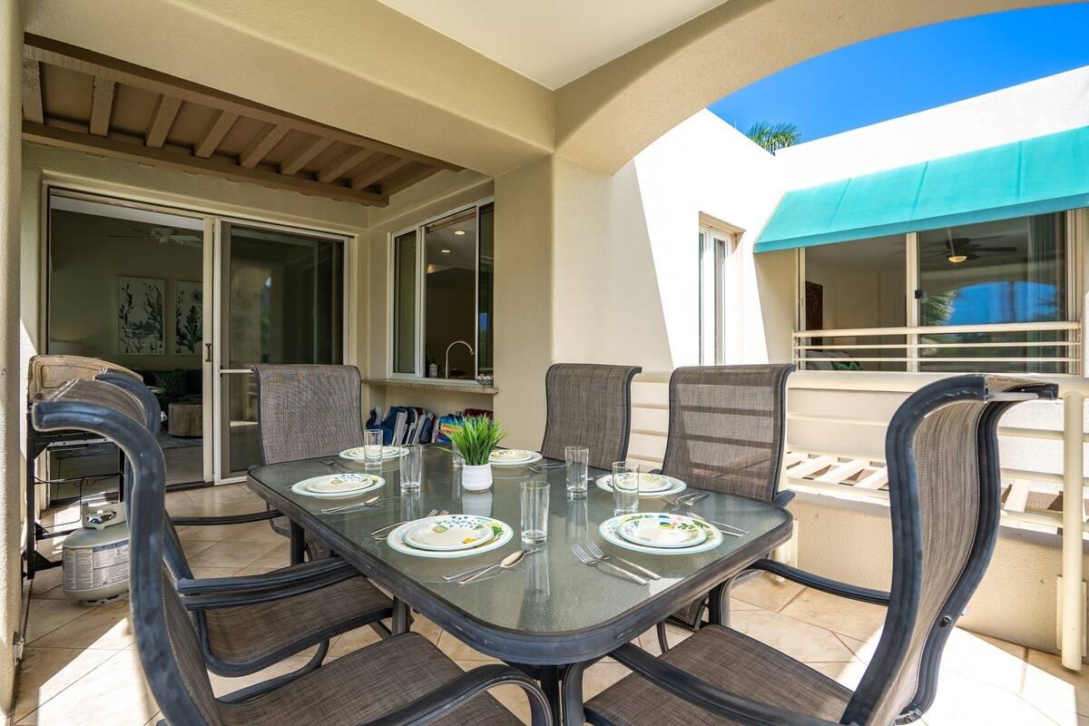 An outdoor patio with a glass table set for dining, surrounded by cushioned chairs.