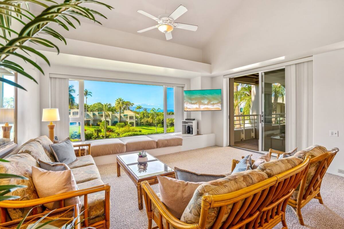 A spacious living room with a large window offering a view of a tropical landscape, a ceiling fan, and a comfortable seating area.