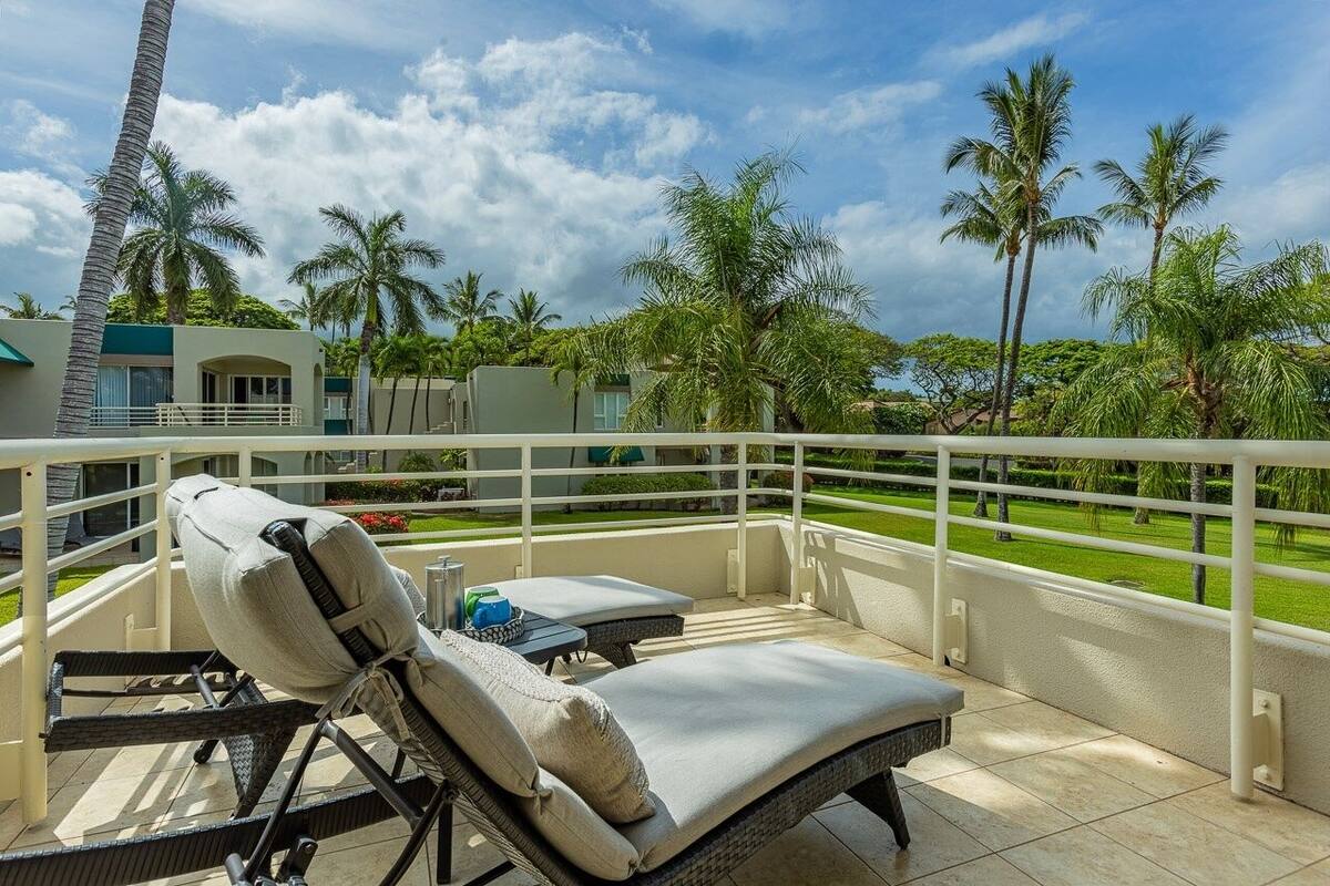 A balcony with two lounge chairs and a small table, surrounded by a white railing and palm trees.