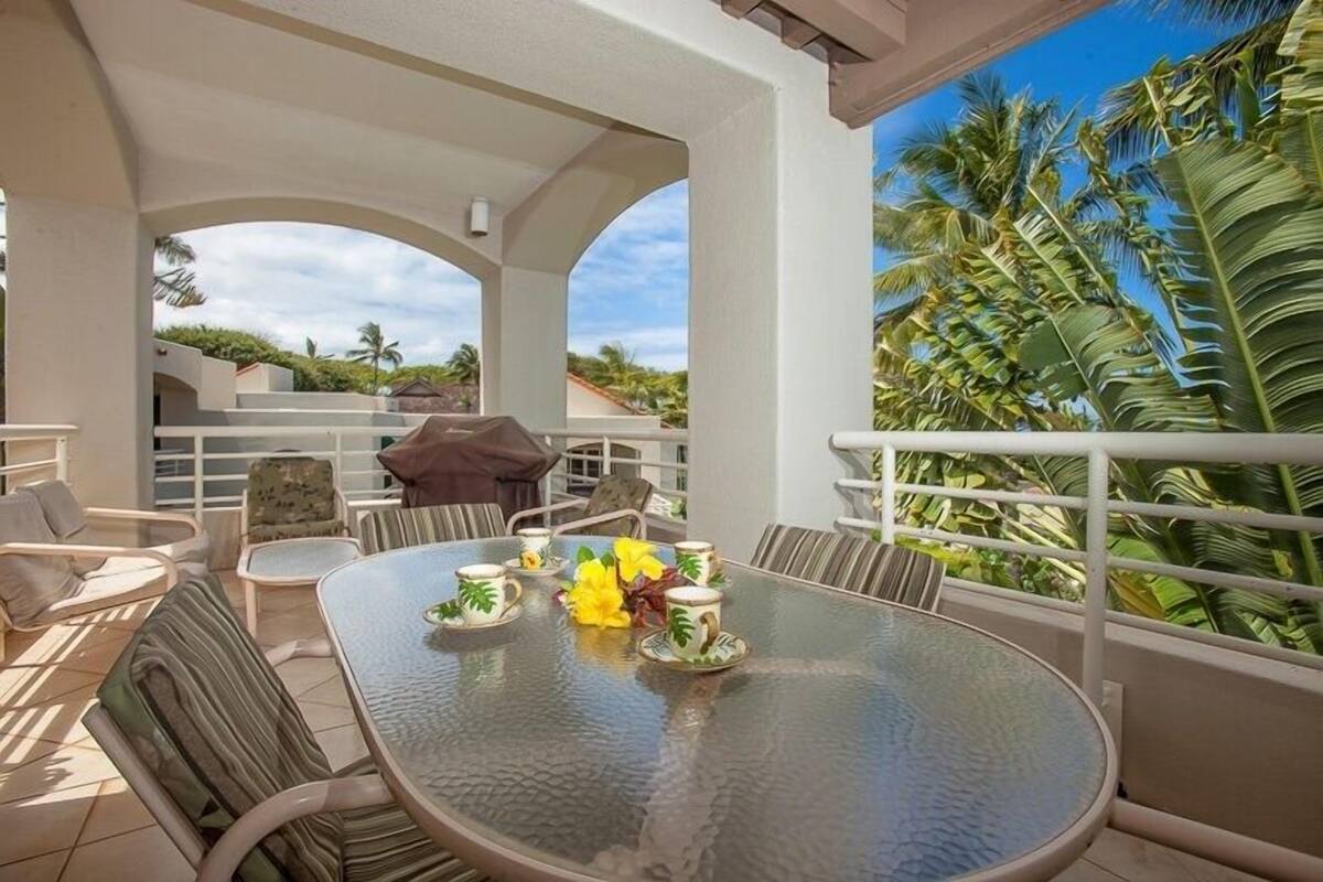 A covered outdoor seating area with a glass table, chairs, and a view of palm trees.