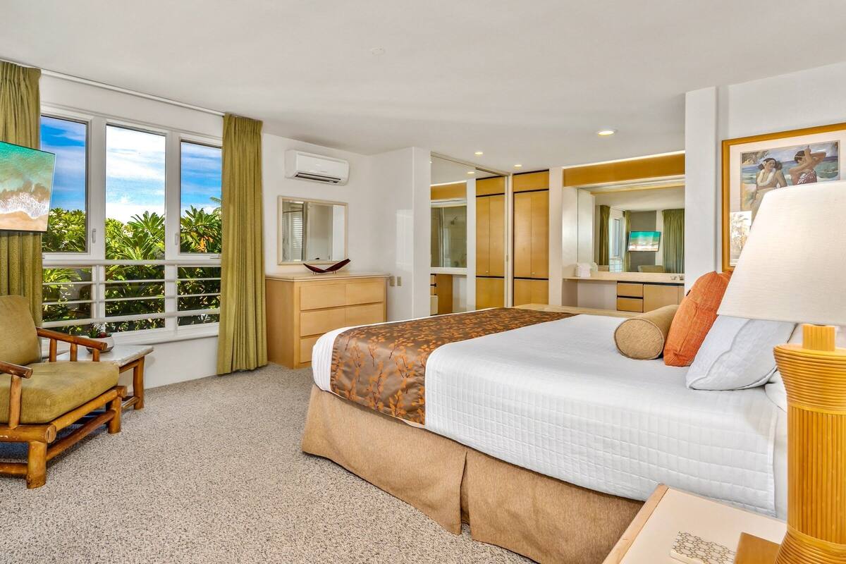 A hotel room with a large bed, a desk, a chair, a TV, and a window with a view of greenery.