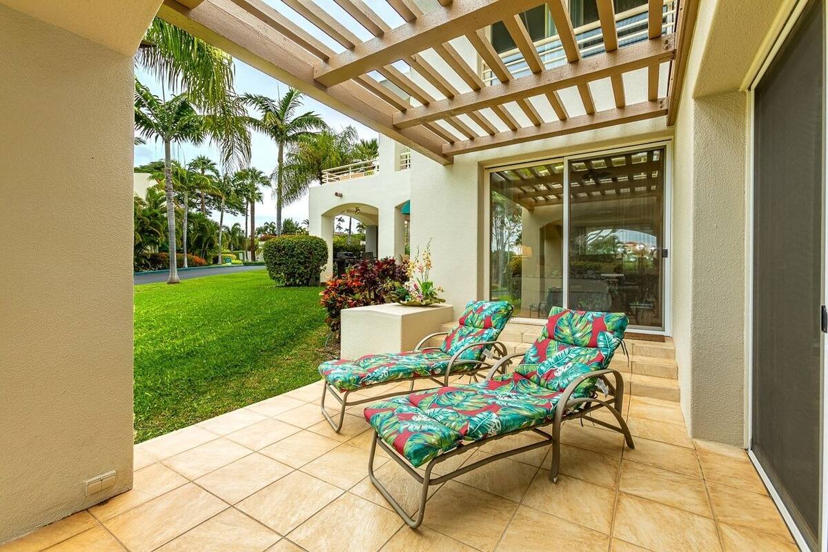 A covered patio with two lounge chairs, a small table, and a view of a well-maintained lawn and palm trees.