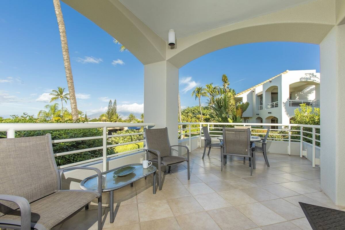 A covered outdoor seating area with chairs and tables, overlooking a landscaped garden and a building with balconies.