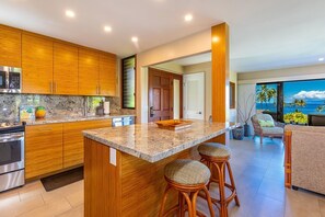 A modern kitchen with wooden cabinets, a granite countertop, and a large island with stools. - Wailea Elua by Coldwell Banker Island Vacations (Kihei)