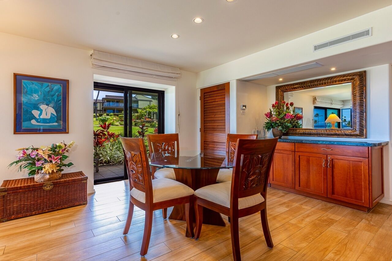 A dining area with a glass table, wooden chairs, a wicker chest, and a large mirror above the counter.