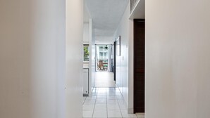 A narrow hallway with white walls and tiled flooring, leading to a room with a window and a door. - Big Island Kona Ali'i by Coldwell Banker Island Vacations (Kailua-Kona)