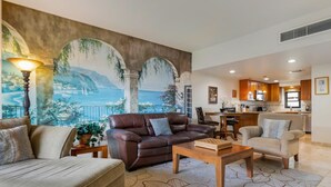 A living room with a large mural, a brown leather sofa, a beige sofa, a wooden coffee table, and a kitchen in the background.