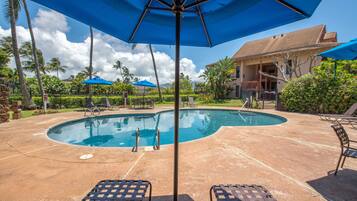 A swimming pool with lounge chairs and umbrellas, surrounded by palm trees and a building with a tiled roof.