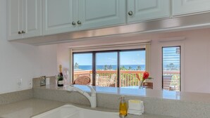 A kitchen with a large window overlooking a balcony with a view of the ocean. - Kauai Makanui by Coldwell Banker Island Vacations (Koloa)