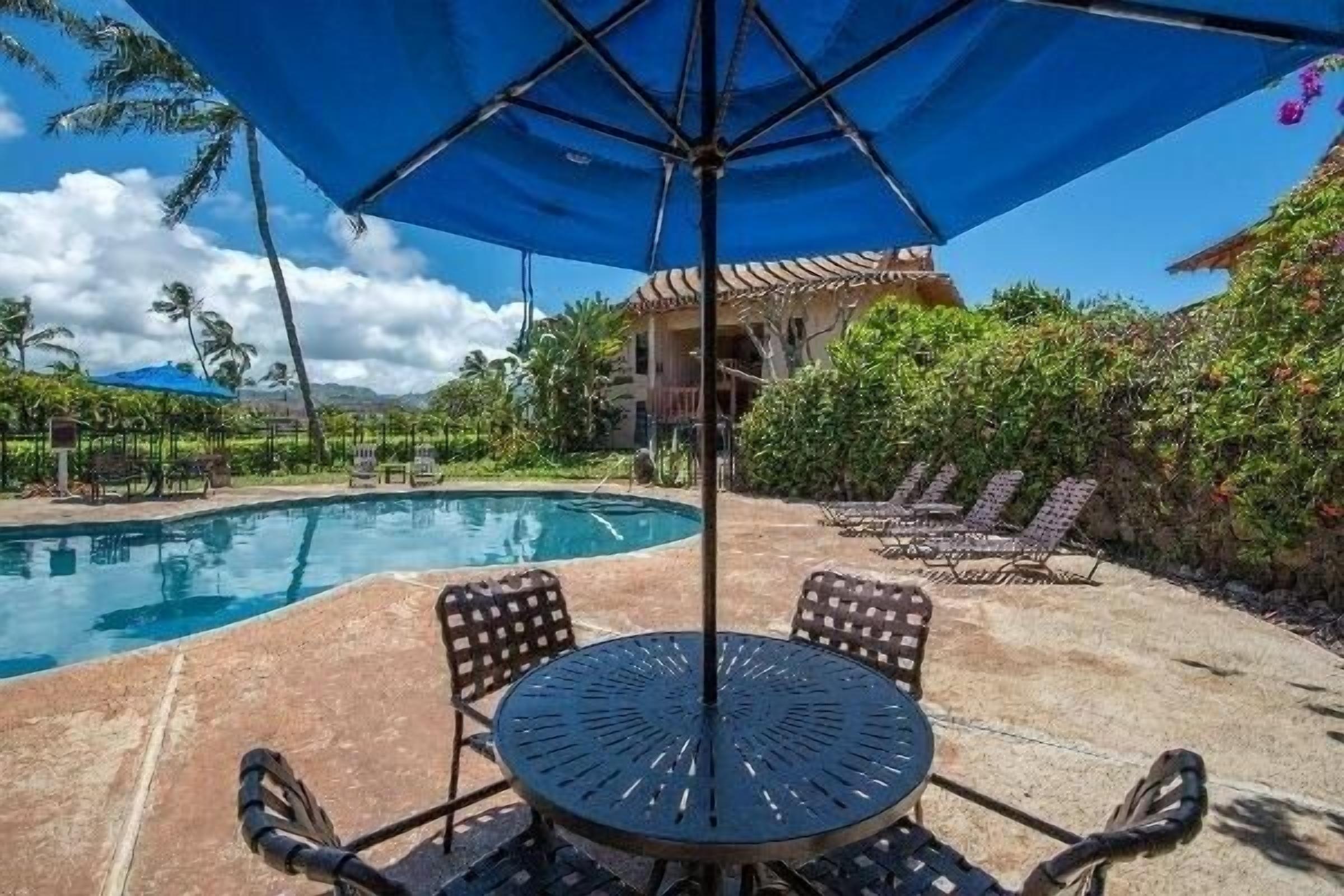 Uma área de piscina com mesa redonda e cadeiras, cercada por espreguiçadeiras e um grande guarda-sol.