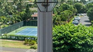 A room with a large window overlooking a tennis court, lush greenery, and distant mountains. - Kauai Makanui by Coldwell Banker Island Vacations (Koloa)
