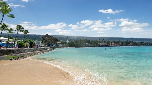 A beach with clear turquoise water, a sandy shore, and a backdrop of palm trees and buildings.