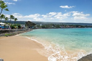 A beach with clear turquoise water, a sandy shore, and a backdrop of palm trees and buildings.