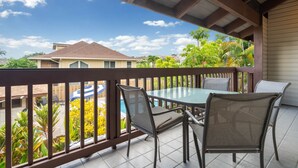 A covered outdoor patio with a glass table and chairs, surrounded by a wooden railing, overlooking a residential area with houses and lush greenery.